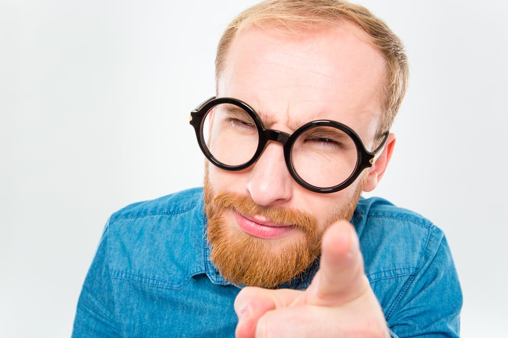 Portrait of suspicious young bearded man in funny round glasses pointing on you isolated over white background-1